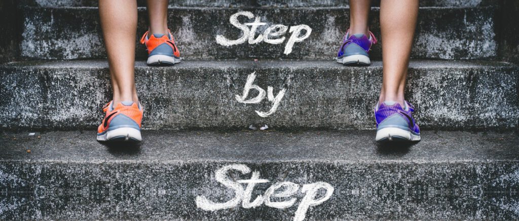 Füße in Sportschuhen, die eine Treppe nach oben gehen. Text auf den Treppen "Step by Step" (Schritt für Schritt)